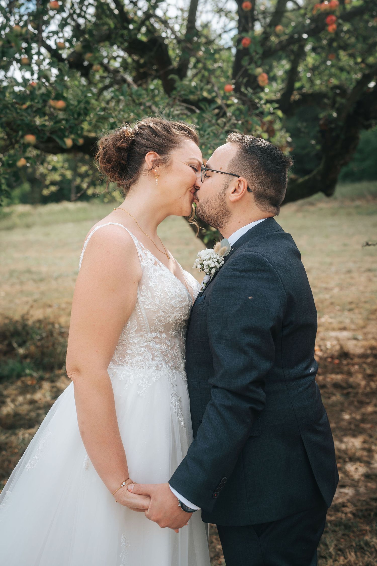 Bride in a lace wedding gown and groom in a navy suit share a kiss beneath an apple tree in a sunny orchard.