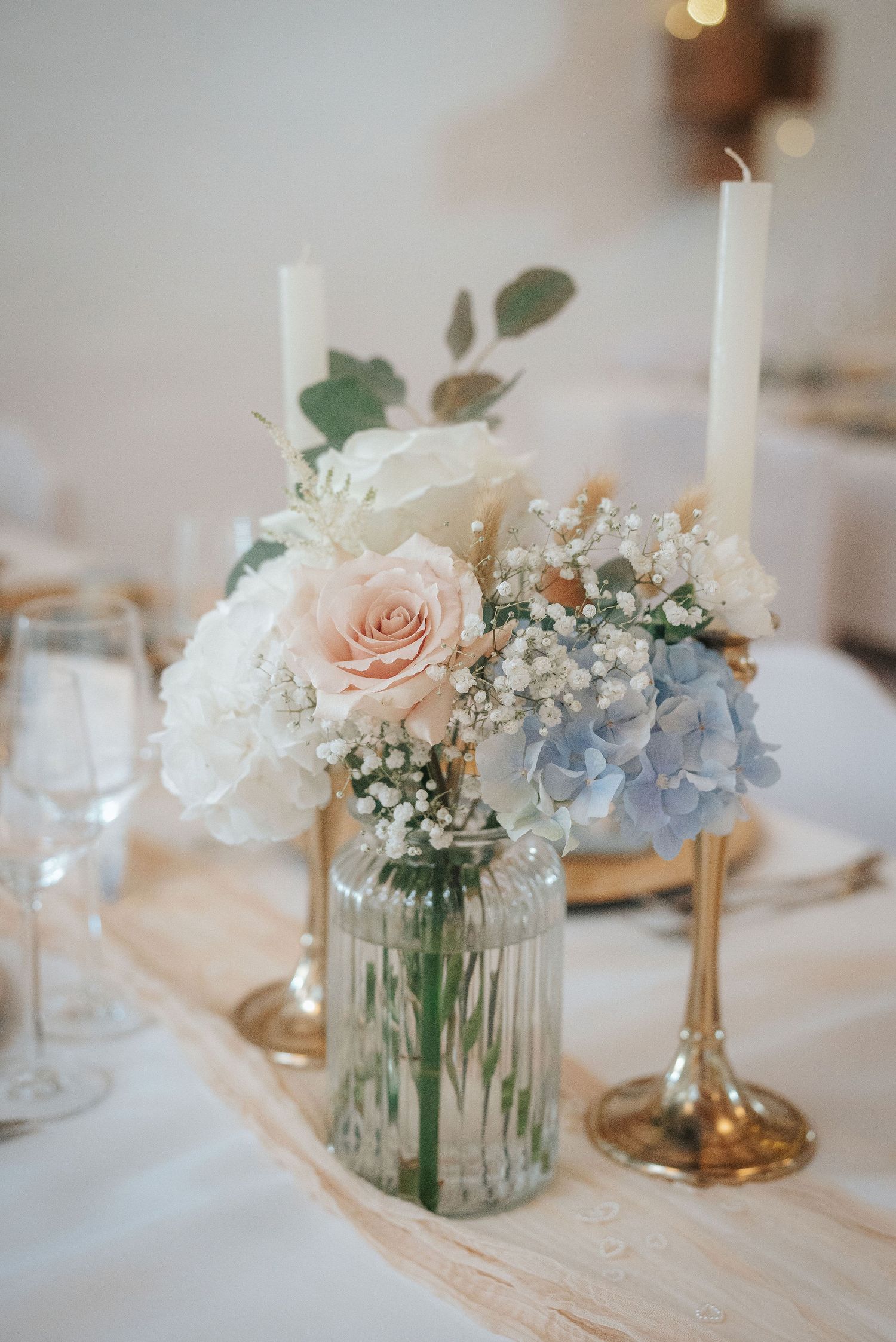 Wedding table centerpiece with blush rose, white hydrangea, baby's breath and blue blooms in a glass jar beside gold candlesticks.