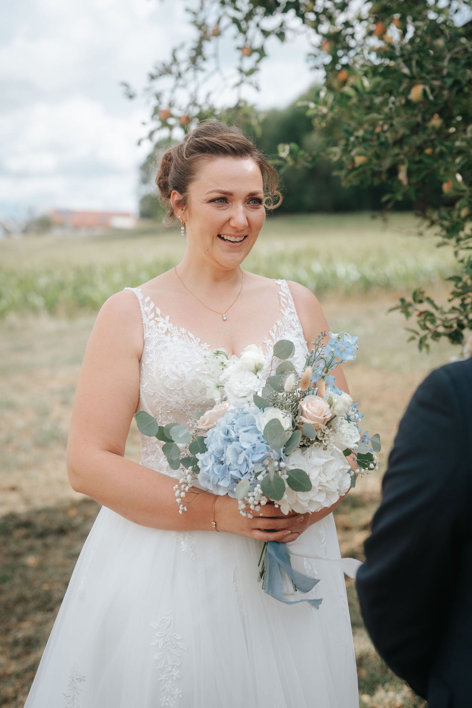 Smiling bride in a lace wedding gown holding a blue and white bouquet with eucalyptus during an outdoor ceremony near an apple tree.