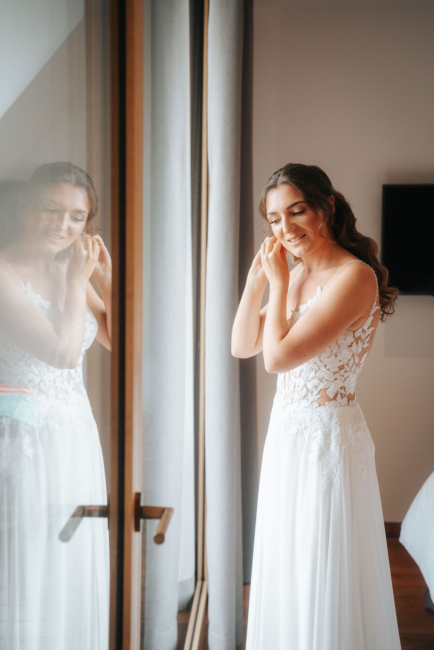 Bride in a white lace wedding gown adjusts her earring beside a tall mirror and window while smiling softly in a bright room.