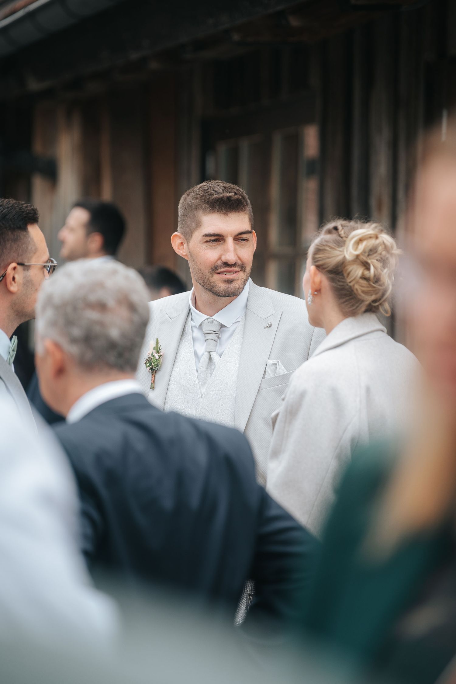 Smiling groom in a light gray suit with a boutonniere chats with wedding guests outside a rustic wooden barn.