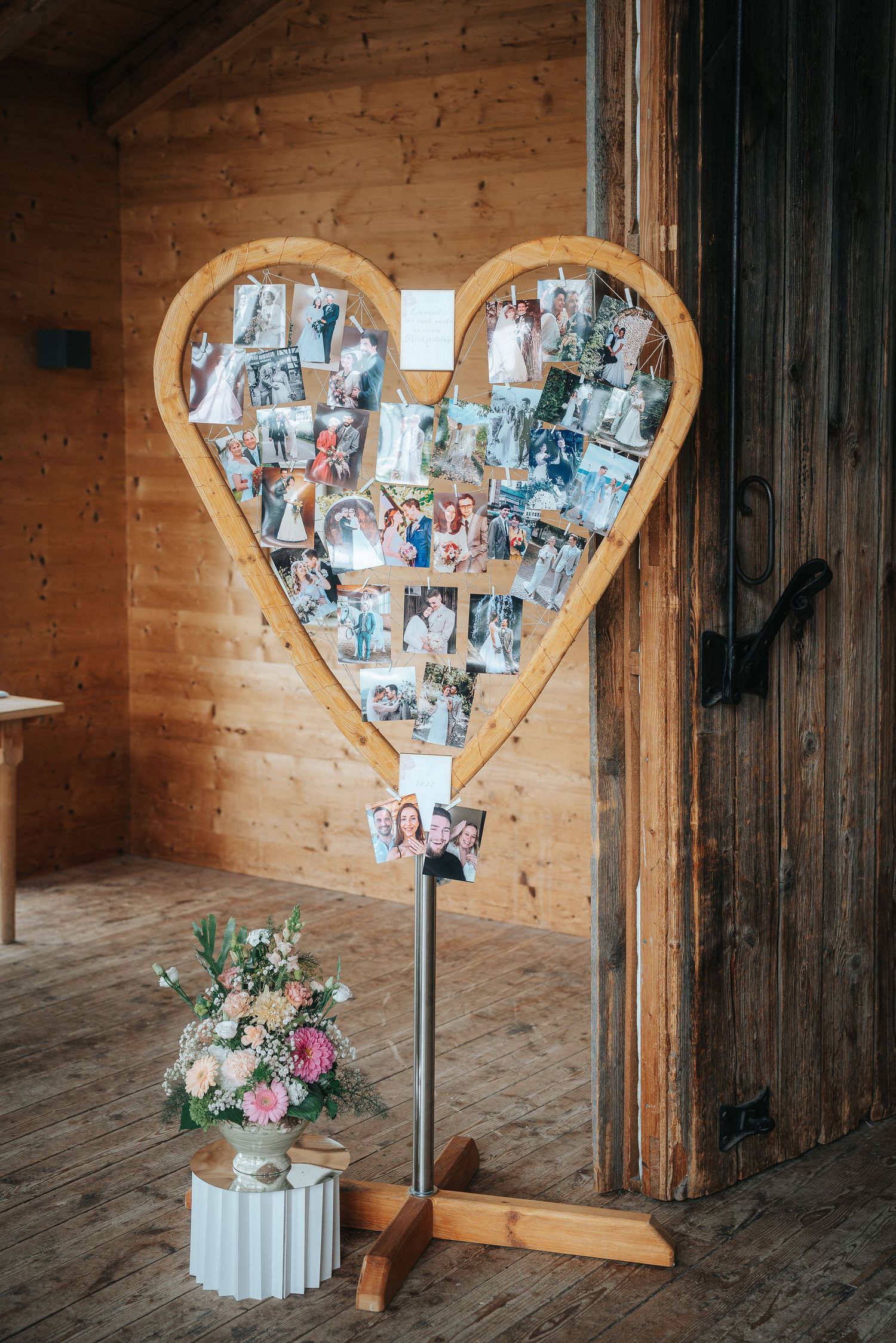Heart-shaped wooden photo display with clipped pictures on a stand beside a pastel flower bouquet in a rustic timber room.