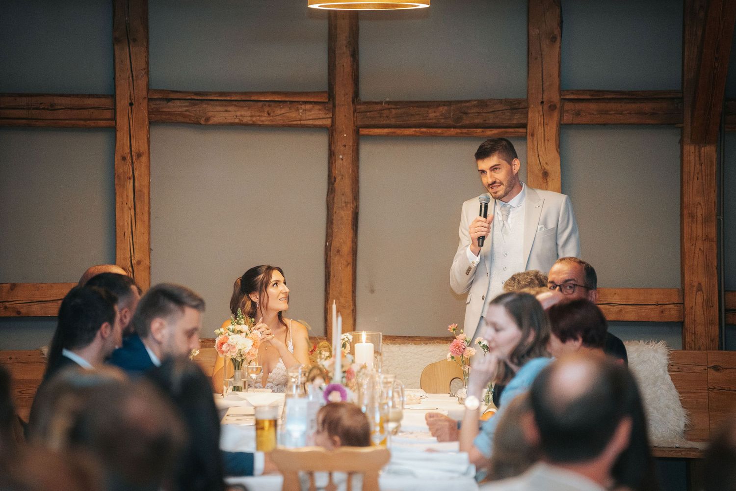Man in a light suit holds a microphone and gives a speech to guests seated at a candlelit table in a rustic timber-framed venue.
