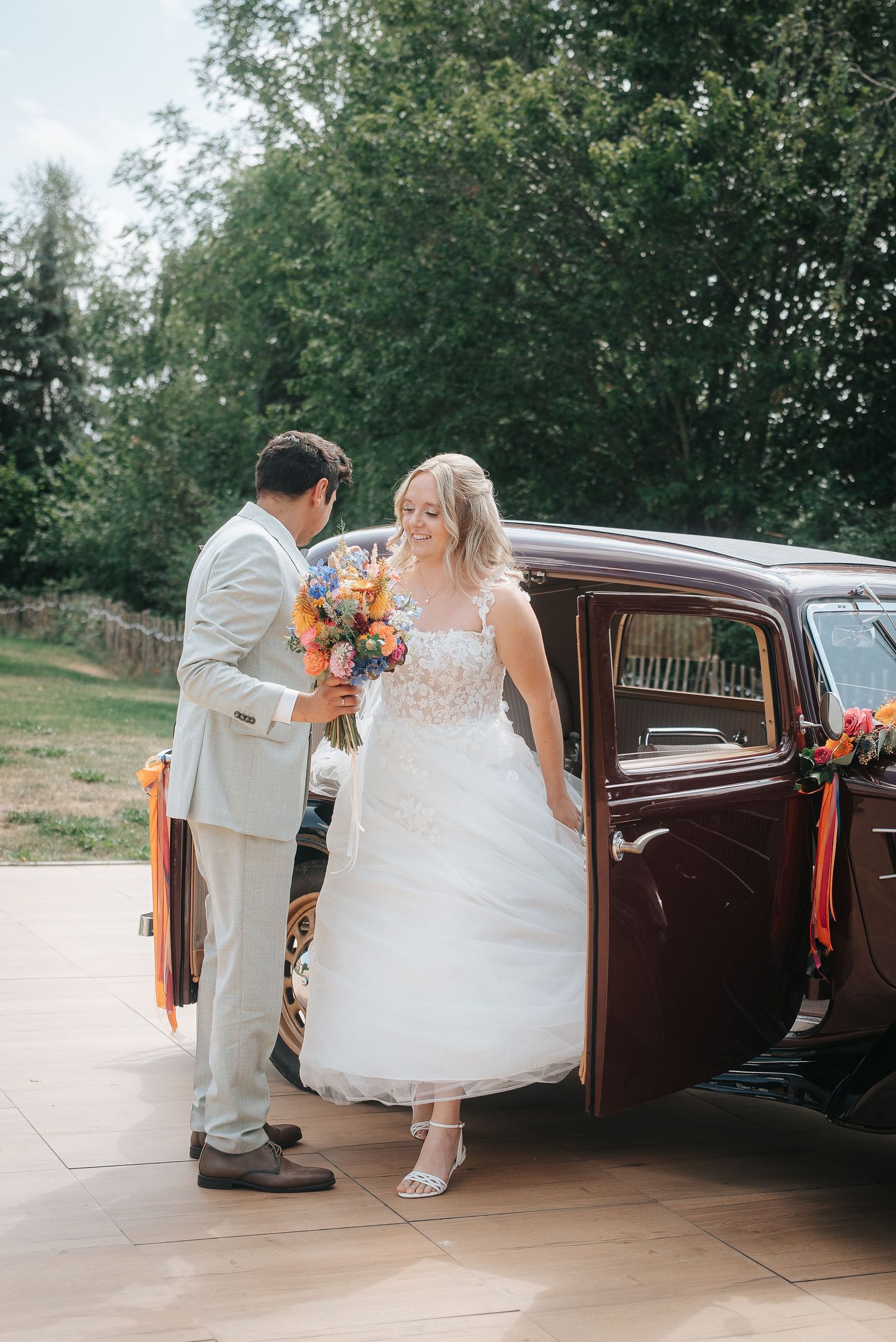 Bride in a white tulle wedding gown steps out of a vintage burgundy car as the groom hands her a colorful bouquet.