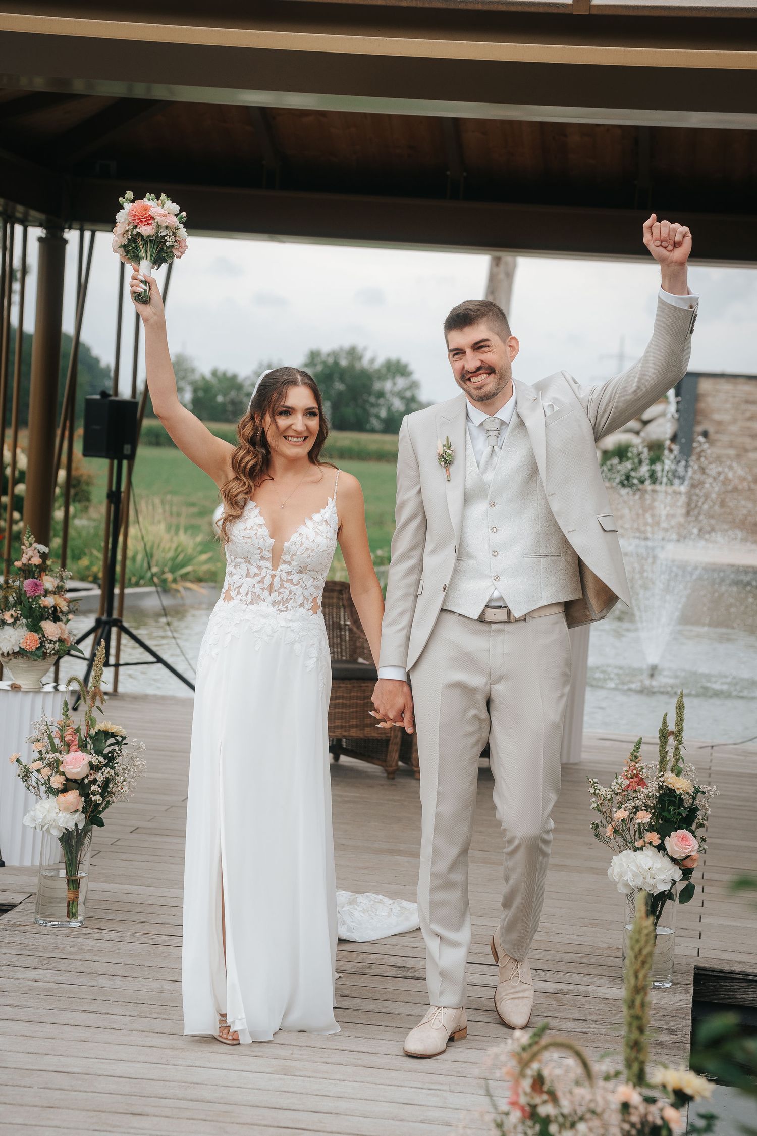 Smiling bride in a lace gown and groom in a beige suit raise their arms in celebration on a wooden deck with flowers and a fountain.