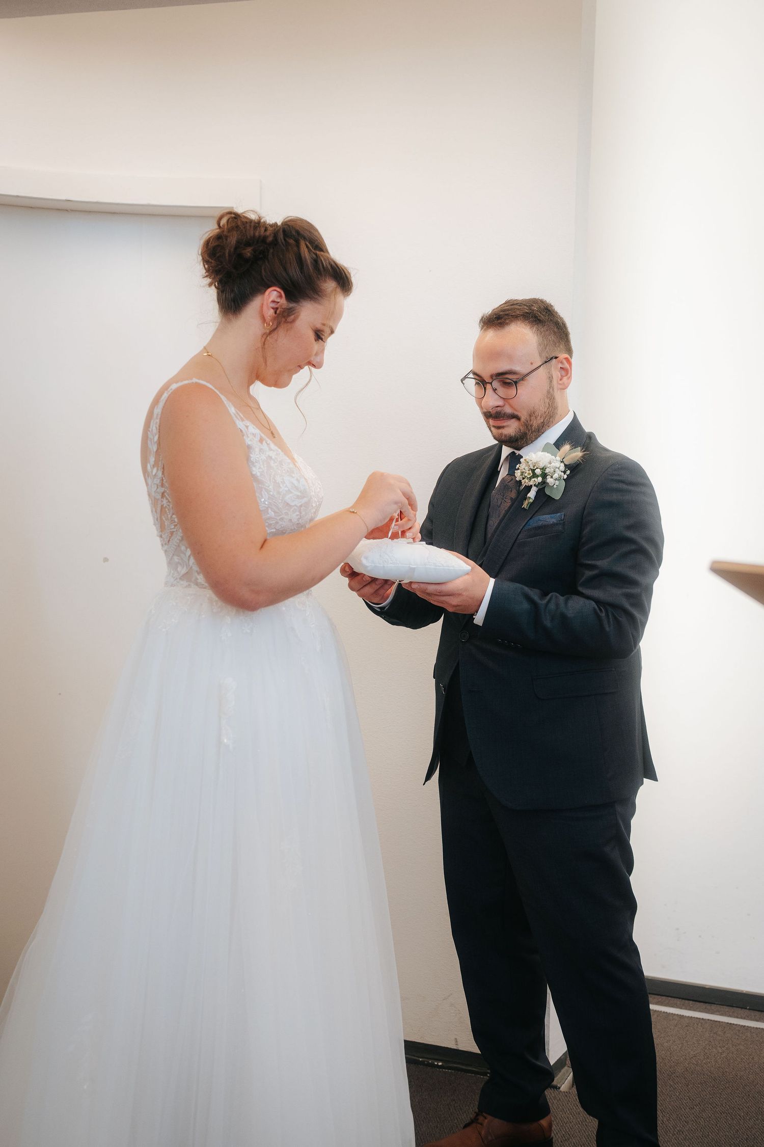 Bride in a white lace and tulle wedding gown takes a ring from a cushion held by a groom in a dark suit during the ceremony.
