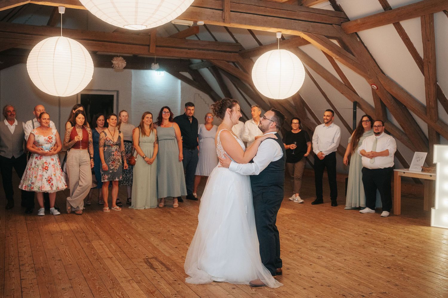 Bride in a white gown and groom share a first dance as wedding guests watch in a rustic barn venue with paper lanterns.