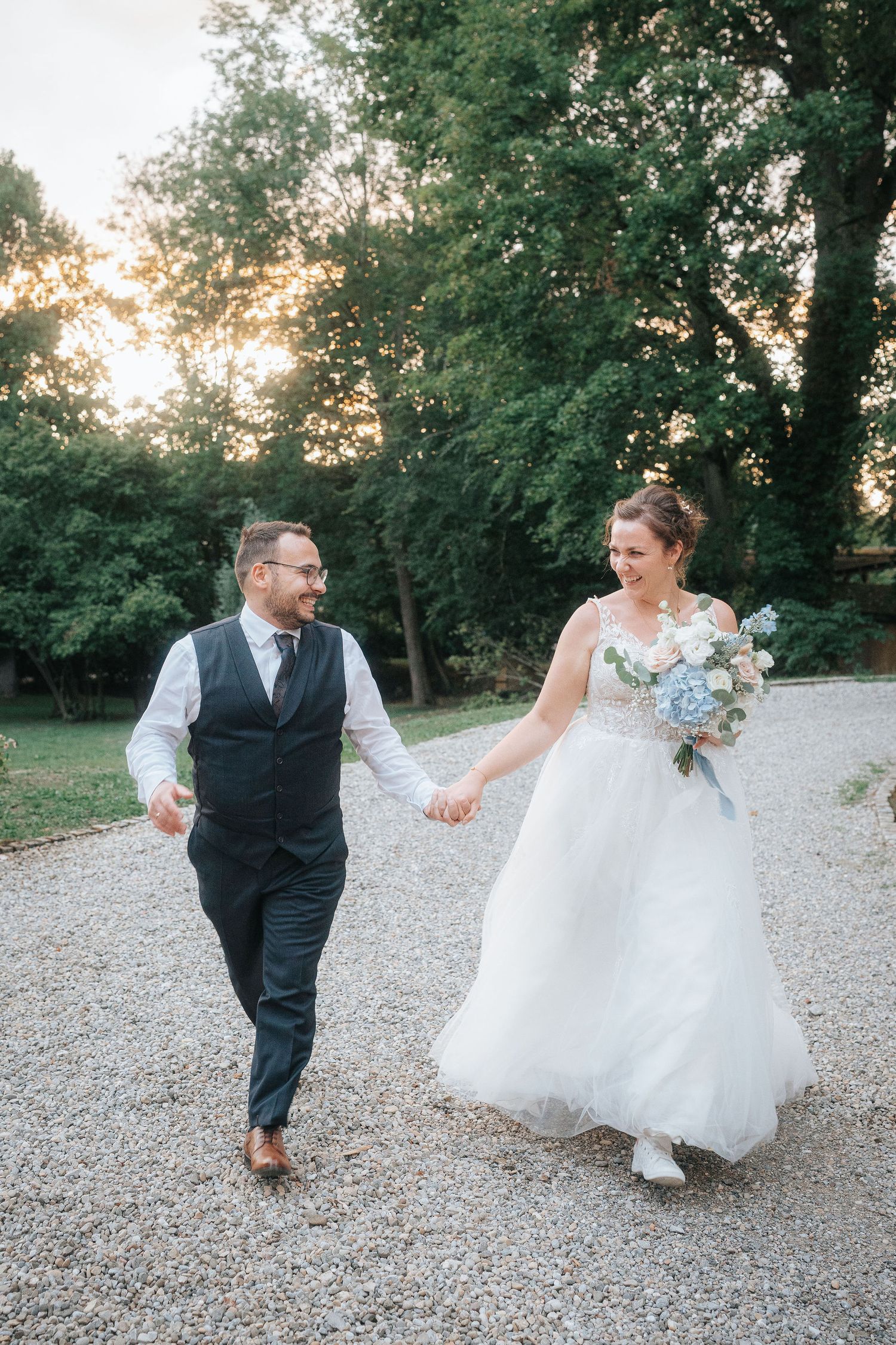 Smiling bride in a white tulle gown holding a blue and white bouquet walks hand in hand with her groom along a gravel path at sunset.