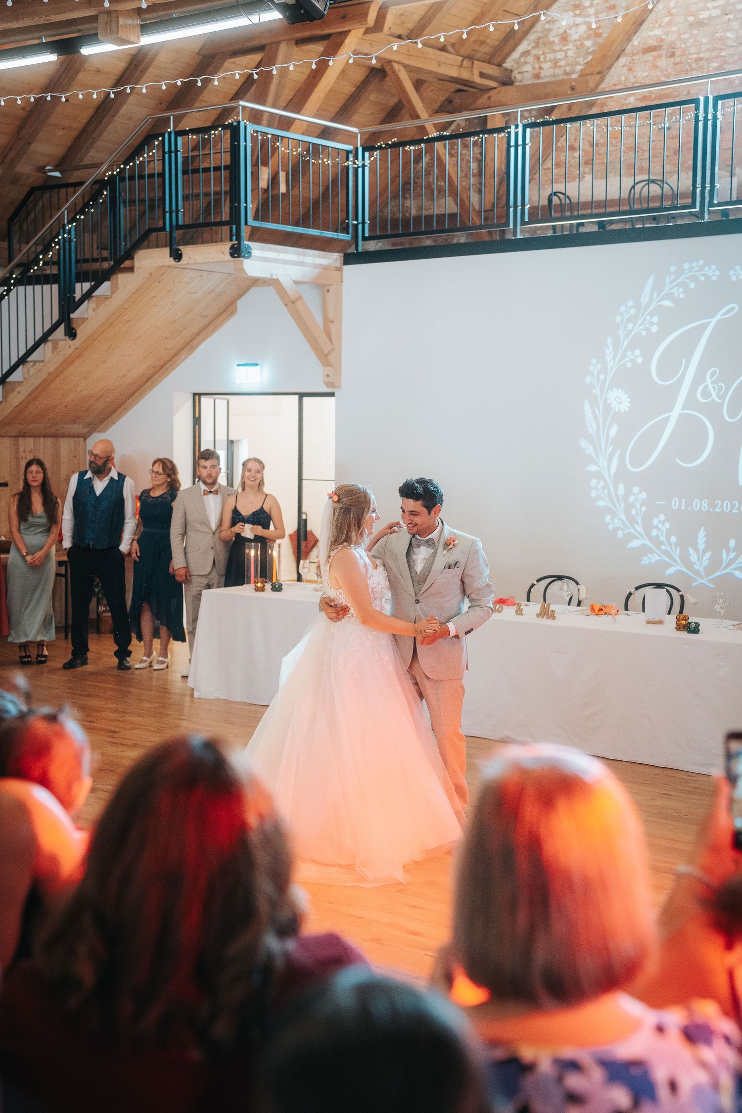 Bride and groom share a first dance in a rustic wedding venue as guests and the wedding party watch beside a monogram projection.