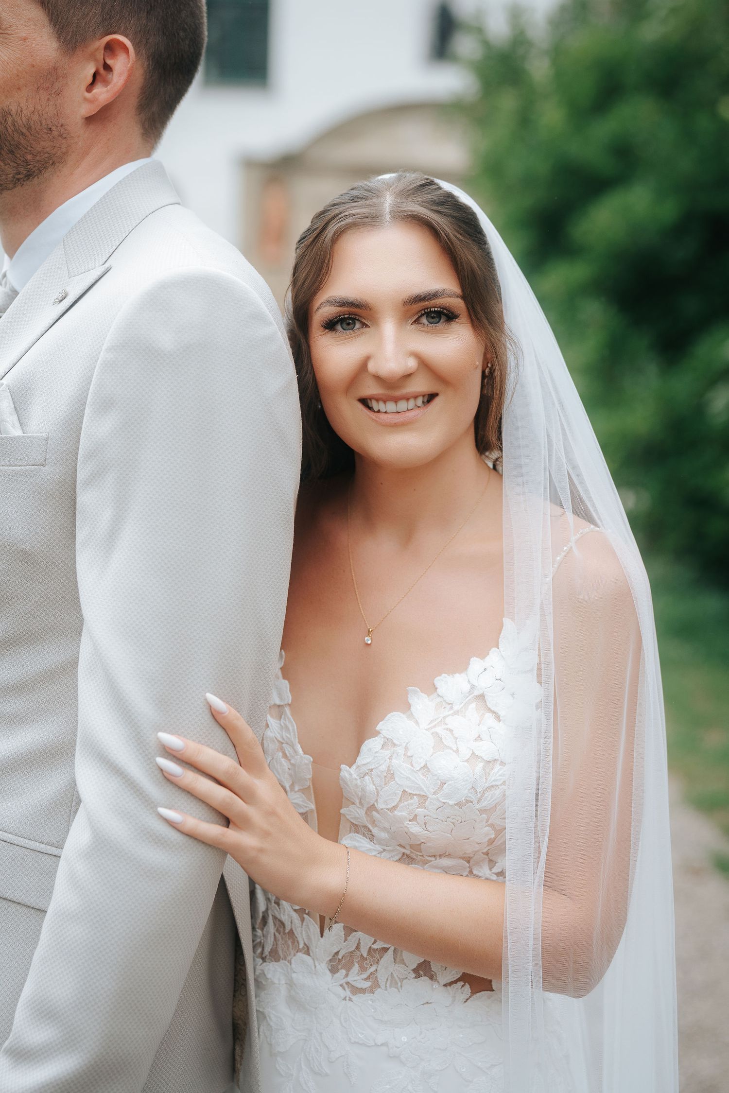Smiling bride in a white lace wedding gown and long veil rests her hand on the shoulder of a groom in a light gray suit outdoors.