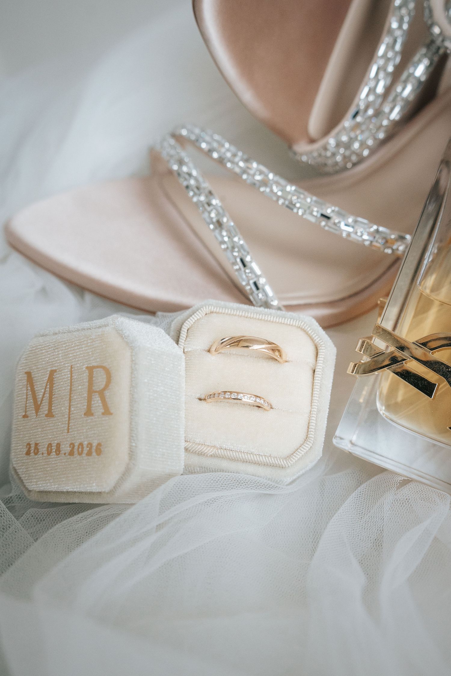 Velvet ring box monogrammed M and R holding two gold wedding bands beside crystal strap sandals and perfume on white tulle.