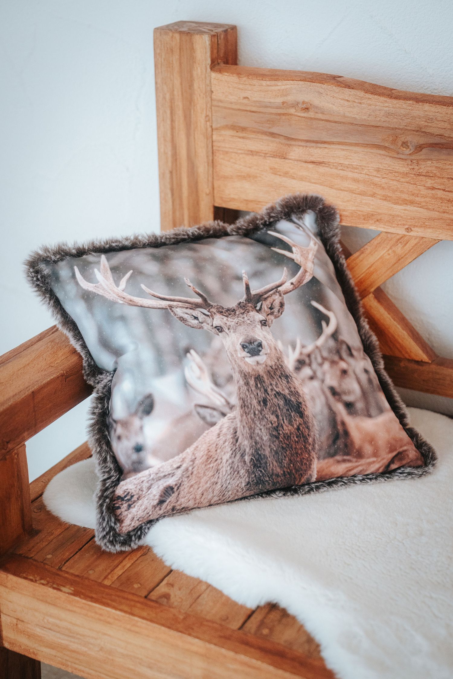 Decorative cushion with a red deer stag photo print and faux fur trim resting on a rustic wooden bench with a white sheepskin throw.