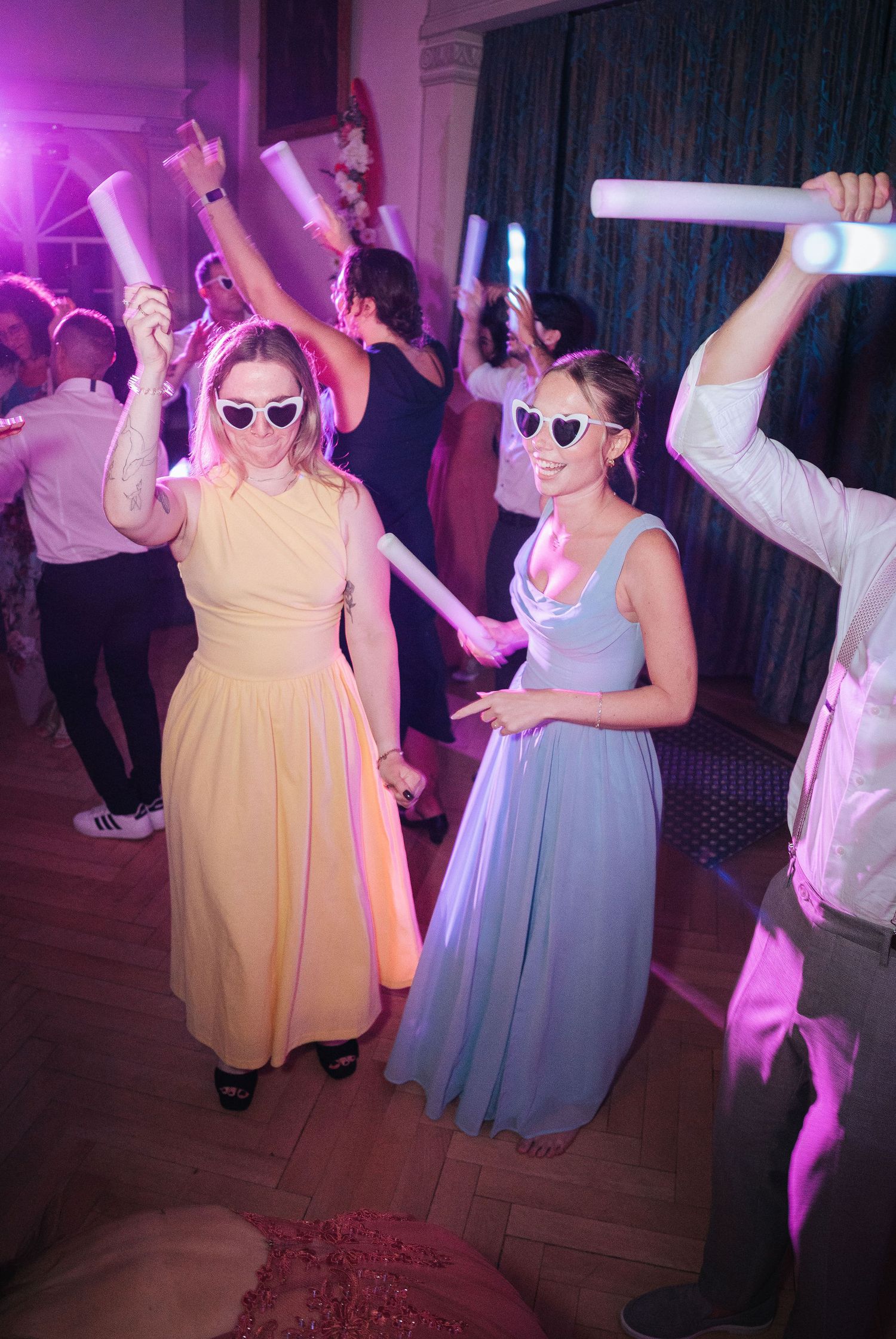 Two women in pastel dresses and sunglasses dance with glowing foam sticks at a lively indoor party under purple lighting.