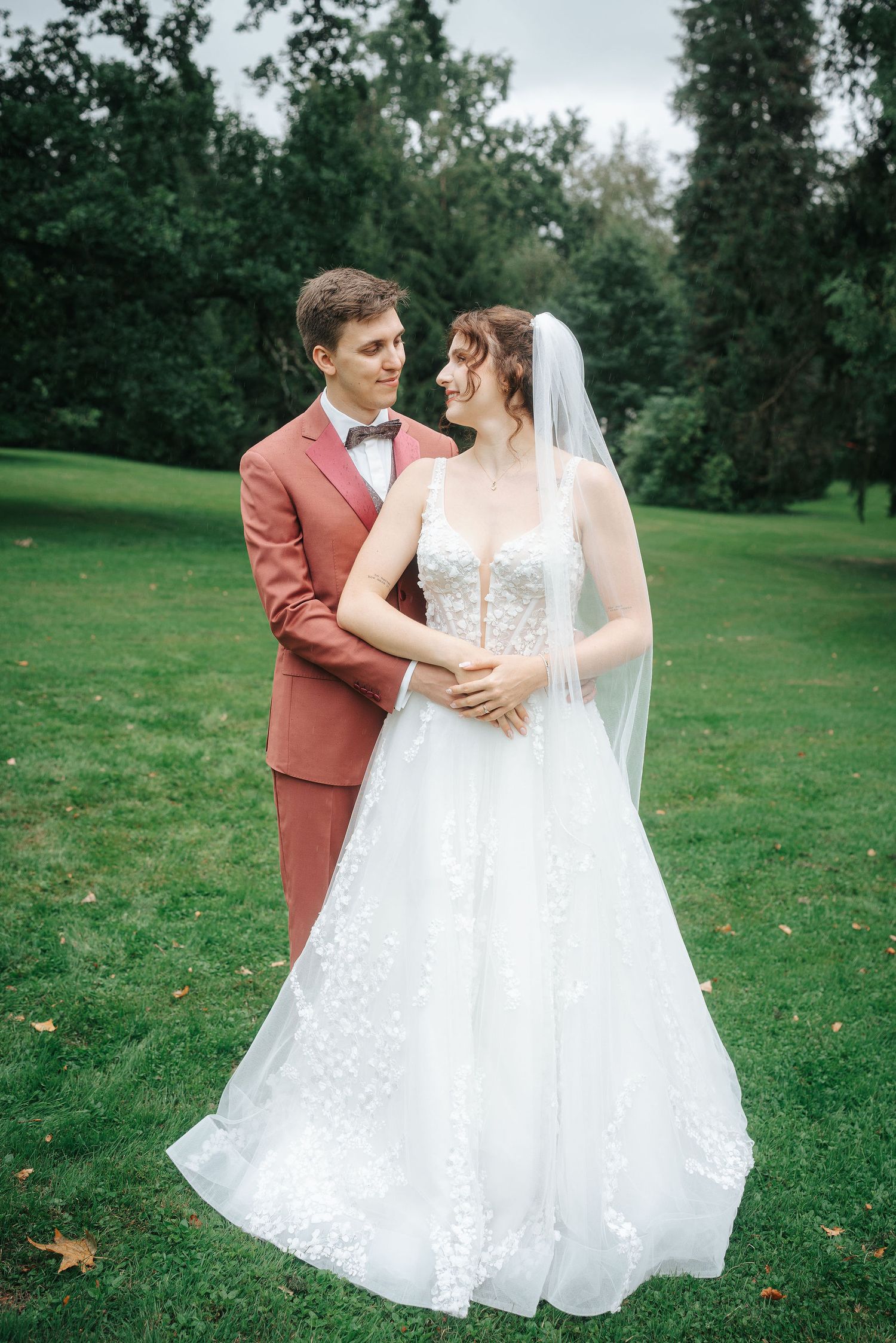 Bride in a lace wedding gown and veil embraces a groom in a rust-colored suit and bow tie on a green lawn surrounded by tall trees.