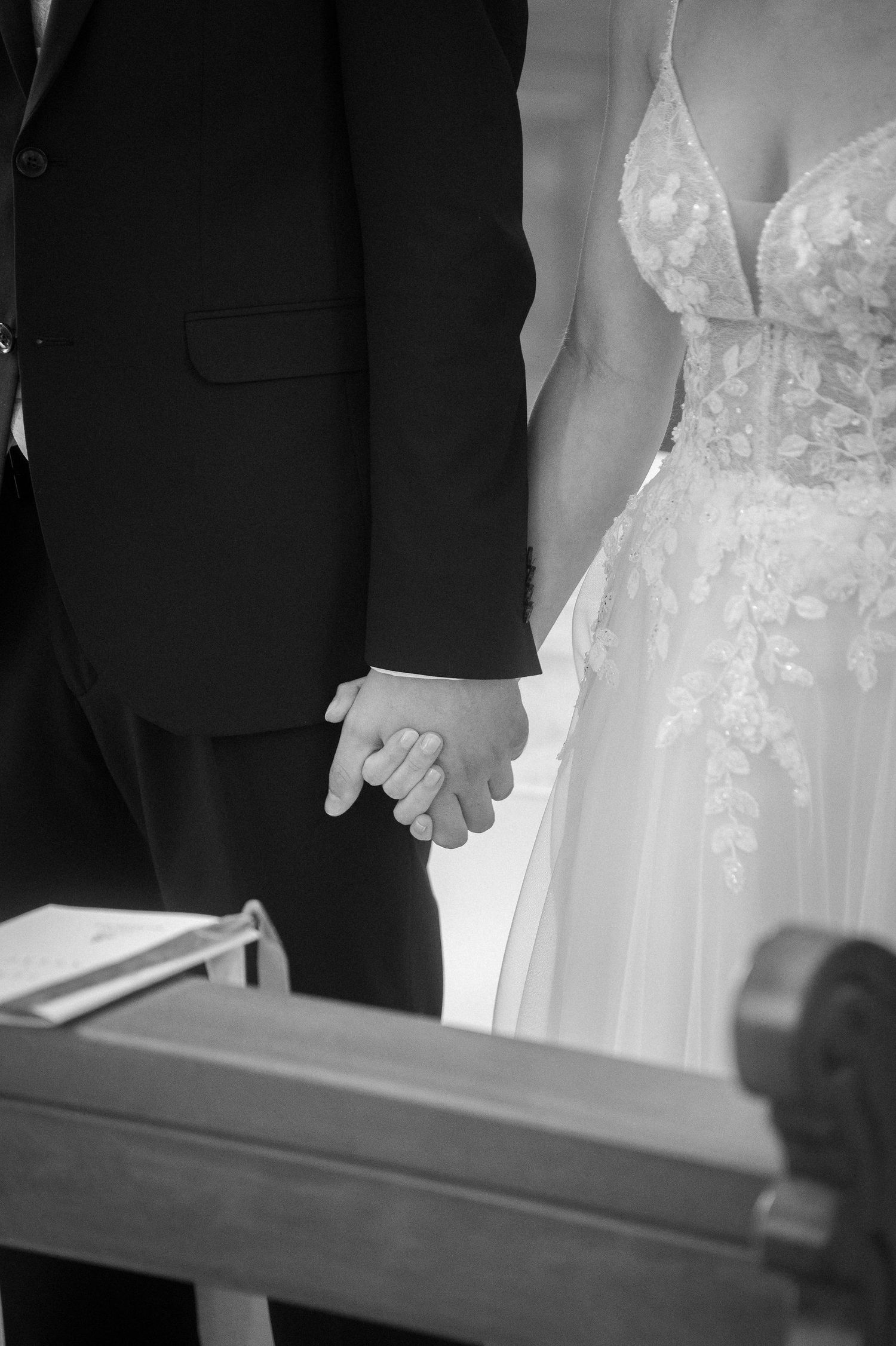 Black and white photo of a bride in a lace wedding gown holding hands with a groom in a dark suit during a church ceremony.