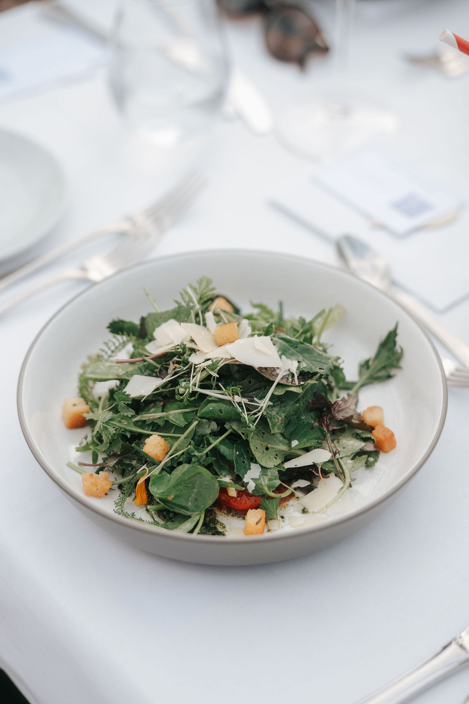 Fresh arugula salad with shaved parmesan, croutons and cherry tomatoes served in a white bowl on an elegant table setting.