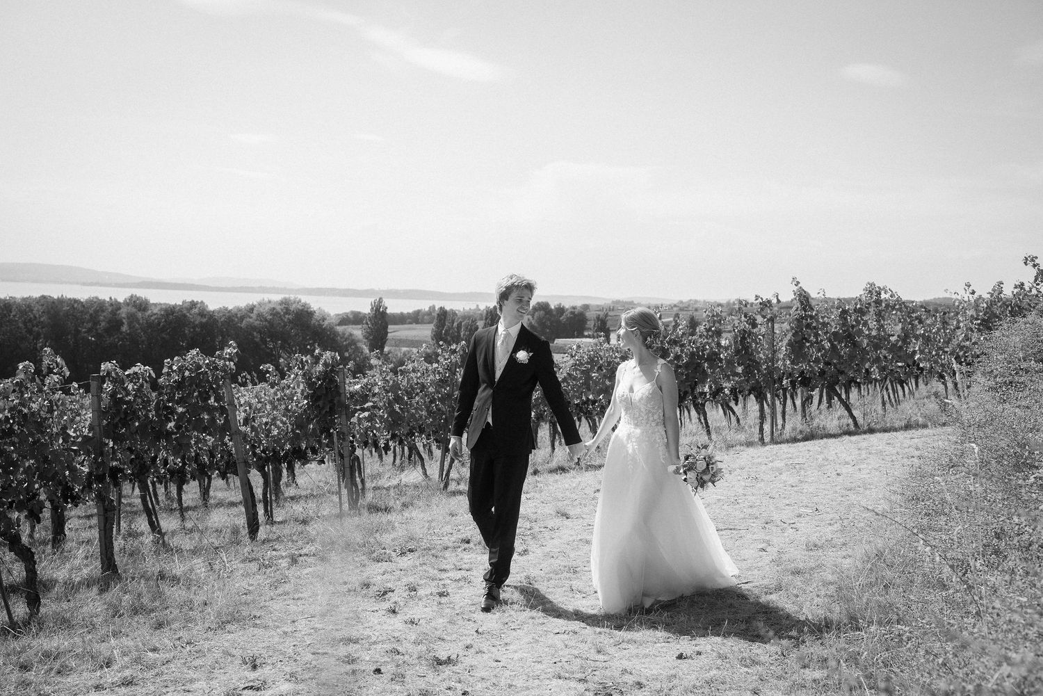 Black and white photo of a bride and groom holding hands and walking along a dirt path through a sunny vineyard.
