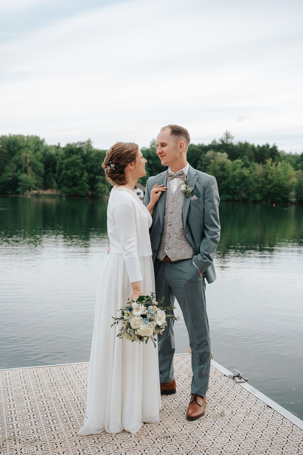 A bride and groom share an intimate moment on a dock by a serene lake, surrounded by trees on a cloudy day.