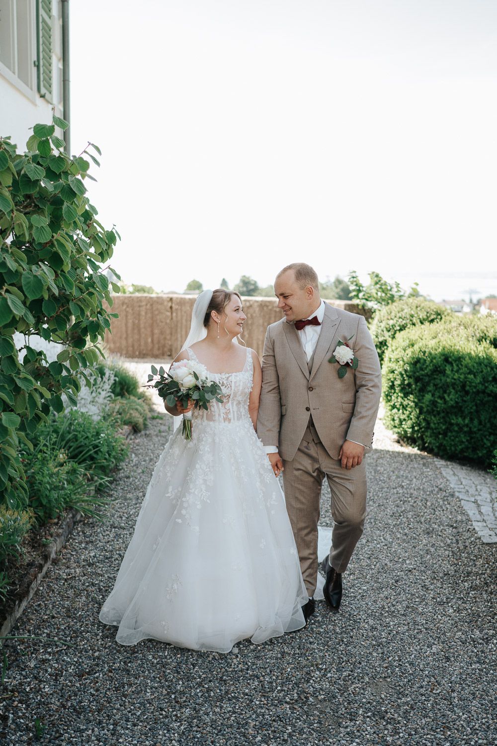 A newlywed couple walks together on a gravel path, with the bride in white and groom in tan suit surrounded by greenery.