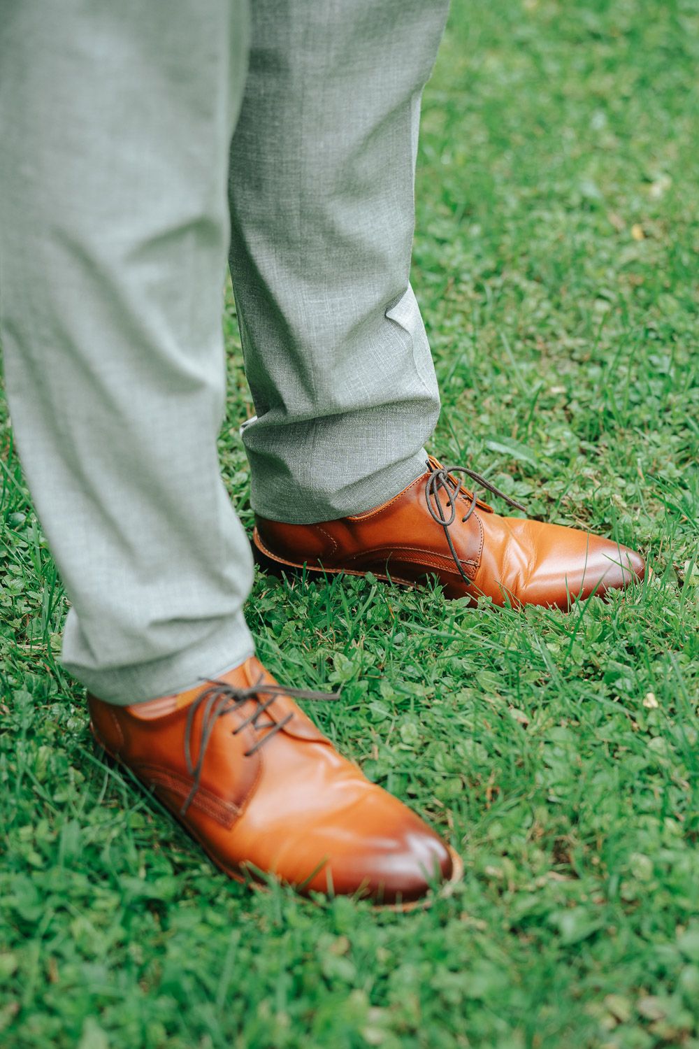 Several angles of brown leather oxford dress shoes worn with light gray suit pants on grass.