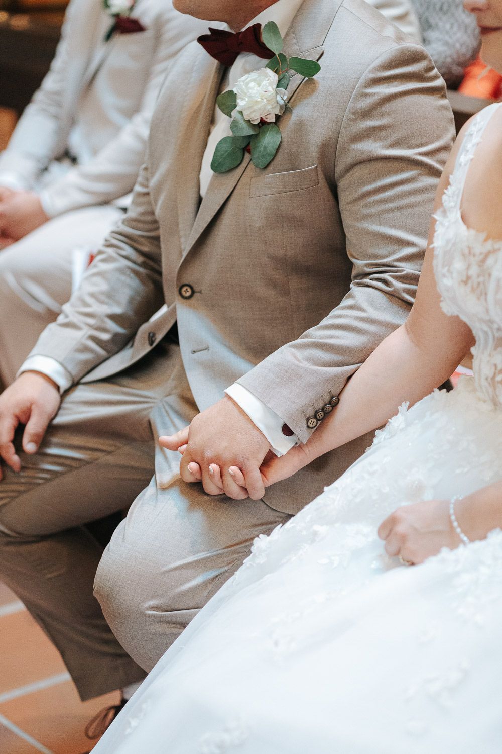 Wedding party members in beige suits and white dresses sit together on bench during outdoor ceremony.
