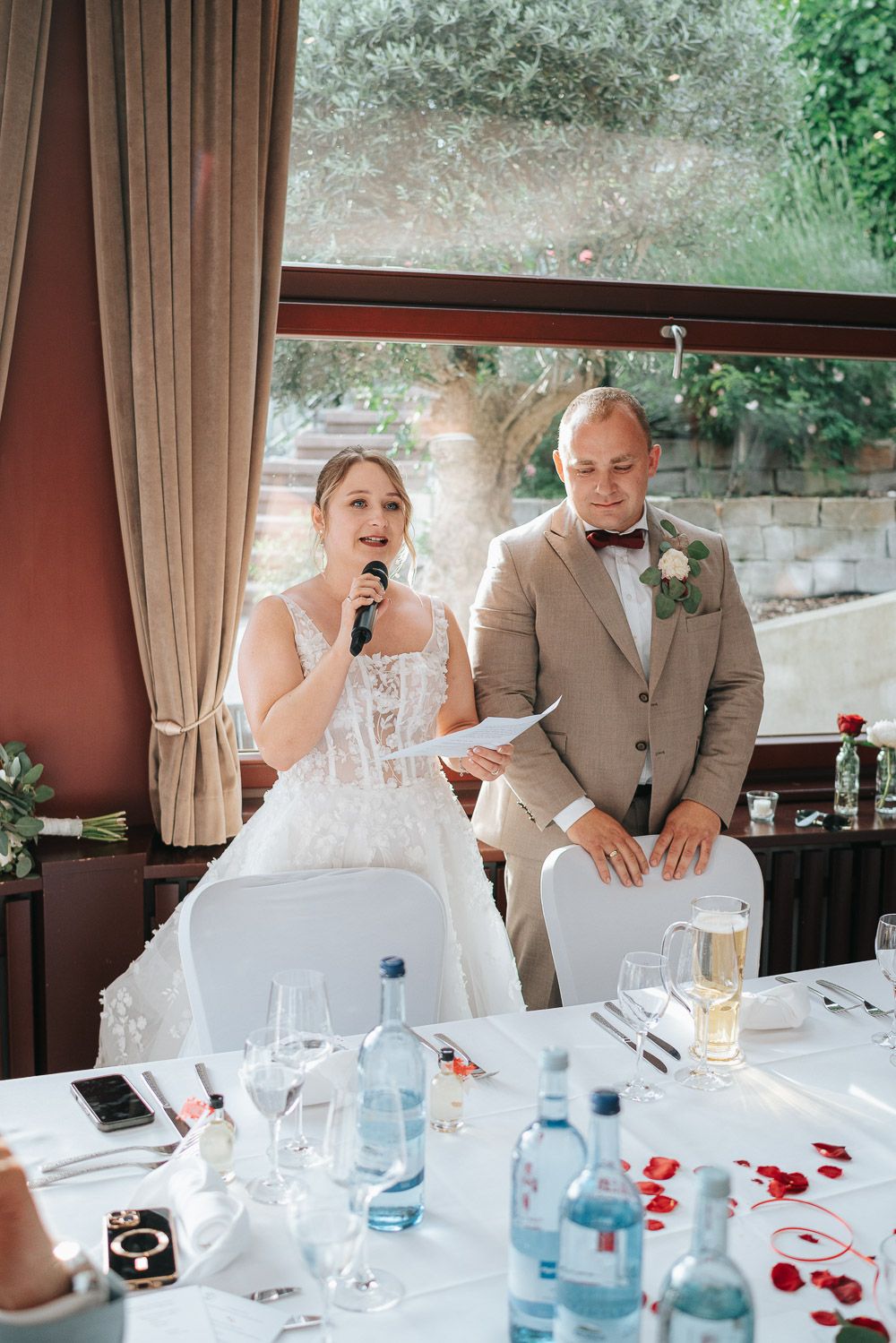 Couple sharing an emotional moment during wedding reception speech at decorated dining table with champagne and rose petals.