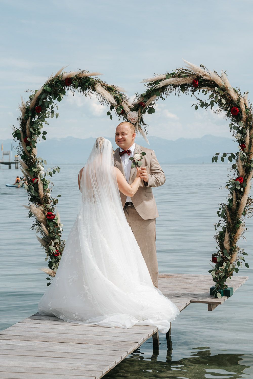 A wedding ceremony takes place on a lakeside dock under a floral heart-shaped arch with mountains in the background.