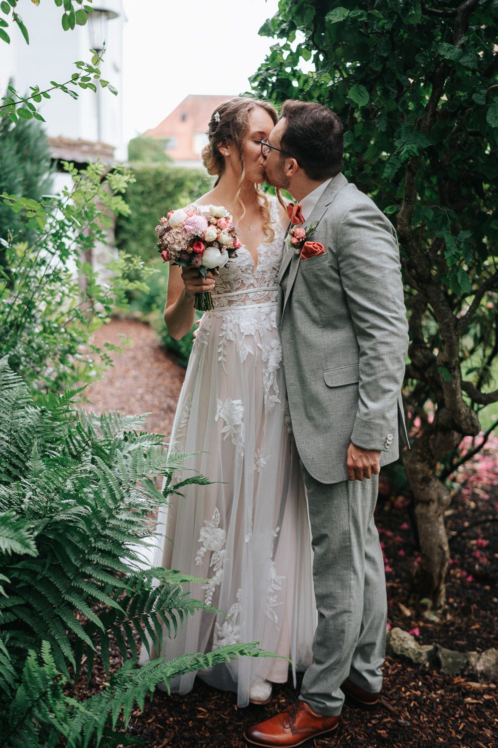 A couple shares a tender moment in a garden setting, with the bride wearing a lace gown and groom in gray suit.