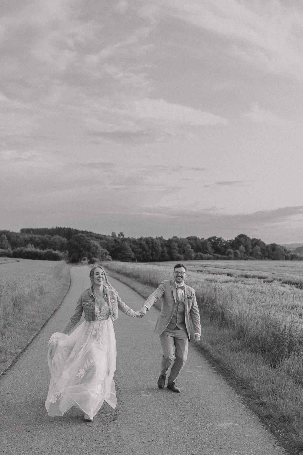 Black and white photo of a couple running together on a country path, with her flowing dress and his suit jacket in motion.