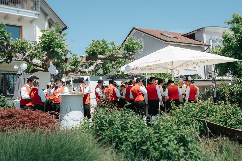 A group in red and white uniforms gathered around outdoor patio tables with white umbrellas in a garden setting.