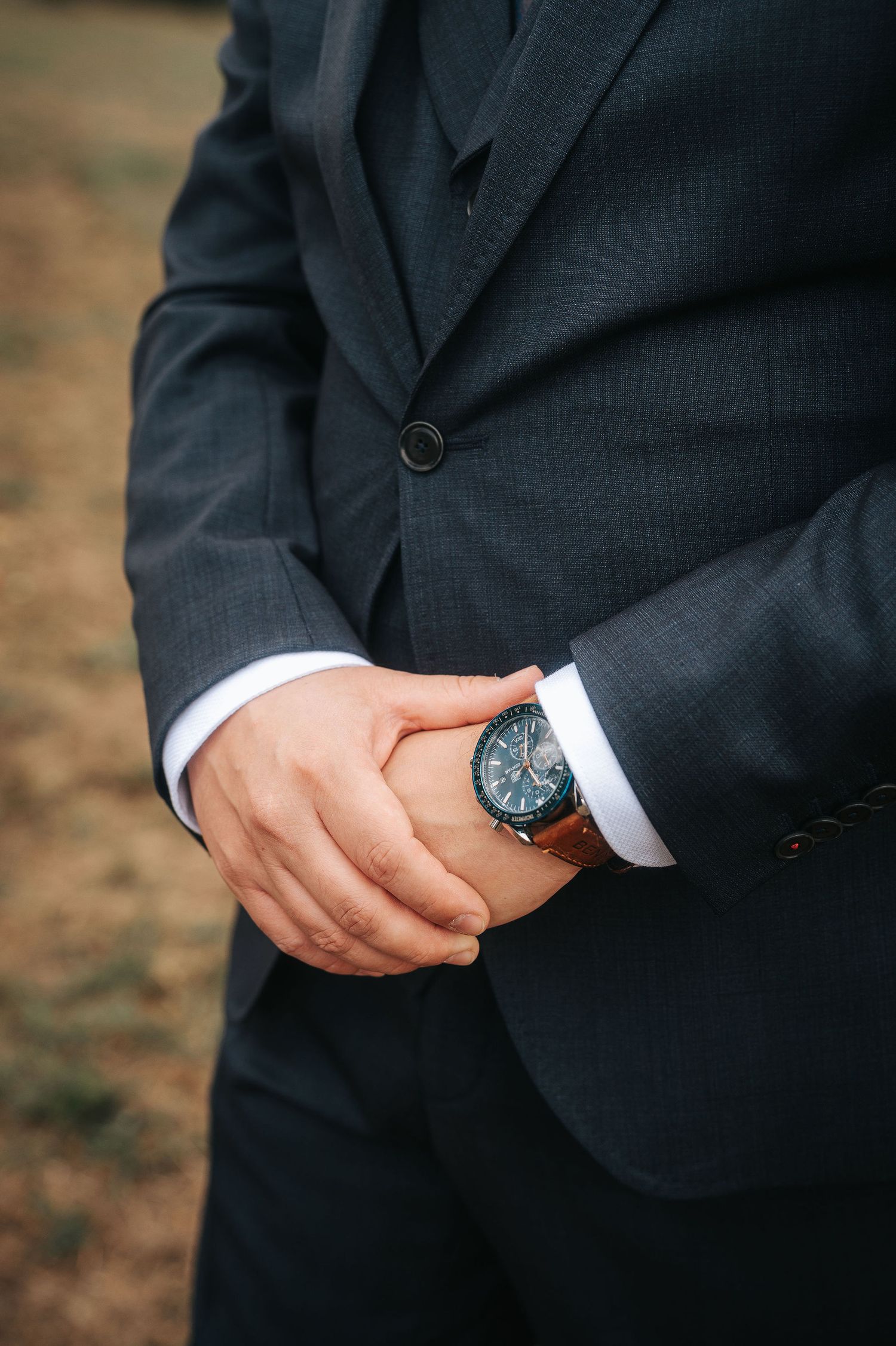Groom in a dark gray suit with clasped hands showing a blue-dial chronograph watch with a brown leather strap outdoors.