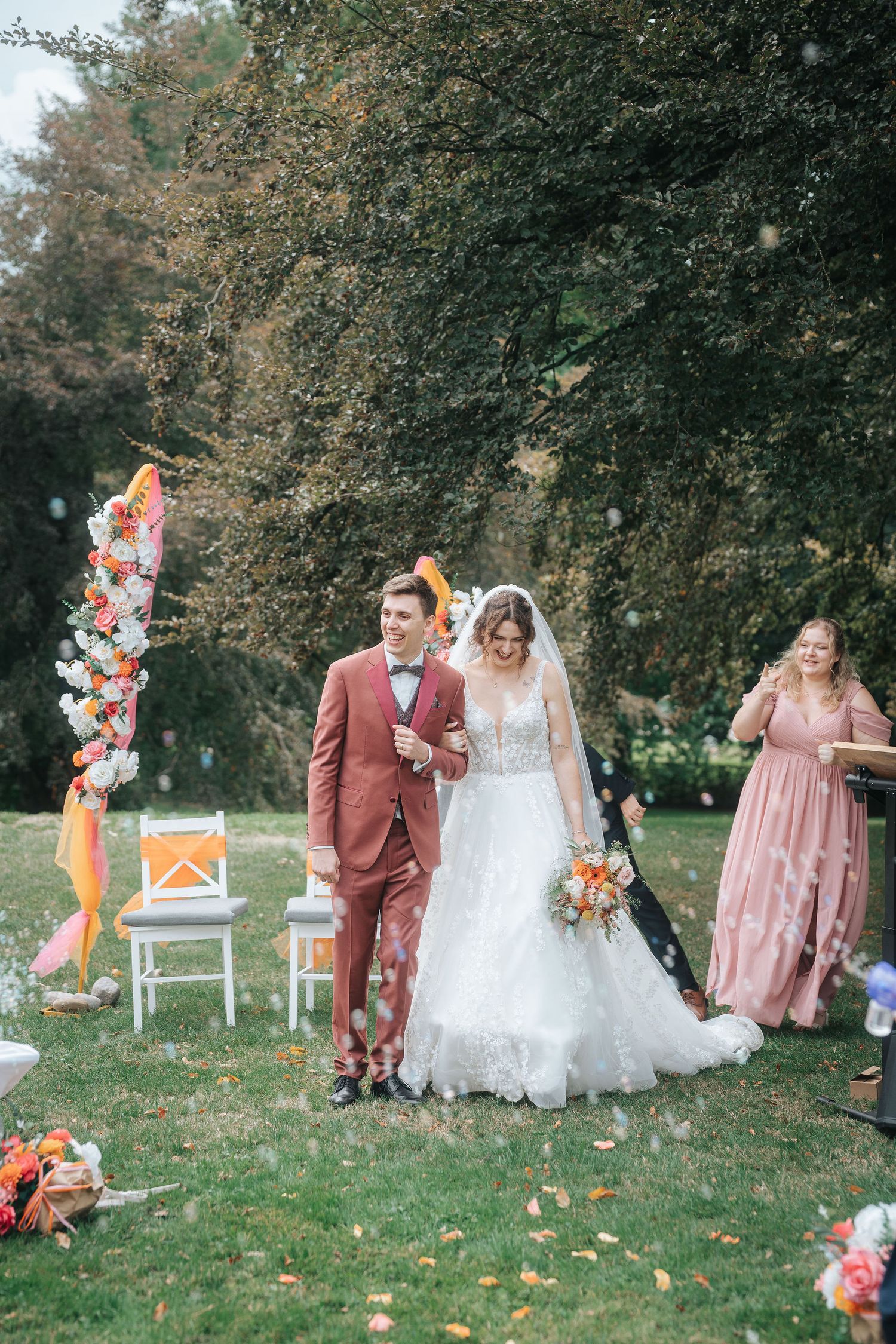 Smiling bride and groom walk down the aisle at an outdoor garden wedding ceremony as guests toss petals and a woman in pink watches.