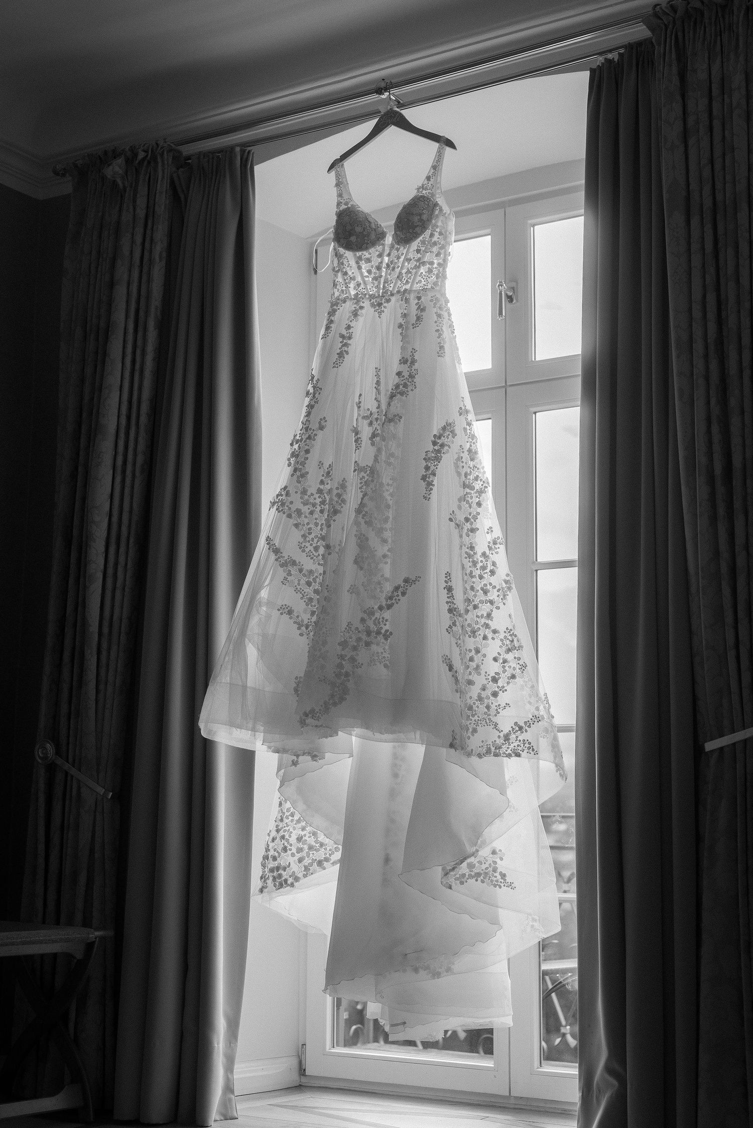 Black and white photo of a lace-embroidered wedding dress with a flowing tulle skirt hanging on a hanger in front of a tall window.