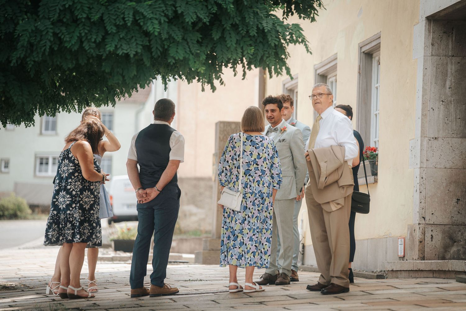 A group of well-dressed adults chats outdoors on a sunny cobblestone square beside a pale building, shaded by leafy green trees.