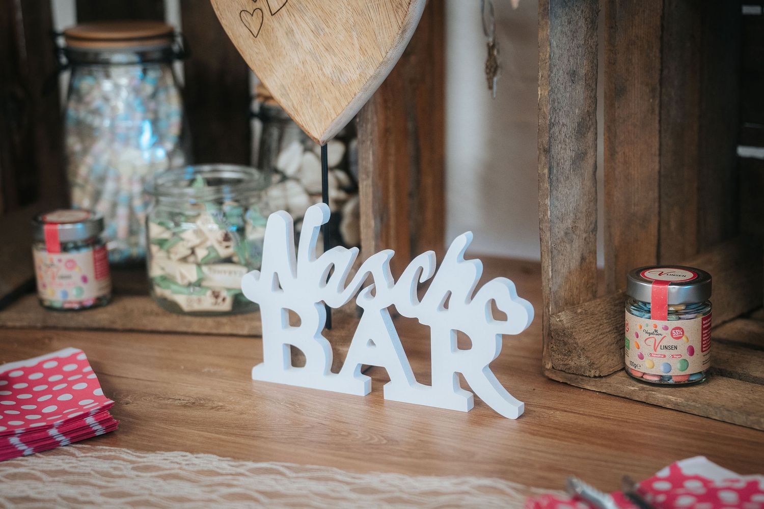 White freestanding Nasch Bar sign on a rustic wooden table surrounded by candy jars and polka dot napkins at a wedding sweets station.