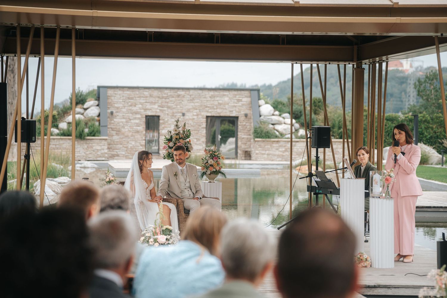 Wedding guests watch an outdoor ceremony as a couple sits beneath a wooden pergola with floral arrangements and a woman in pink officiates.