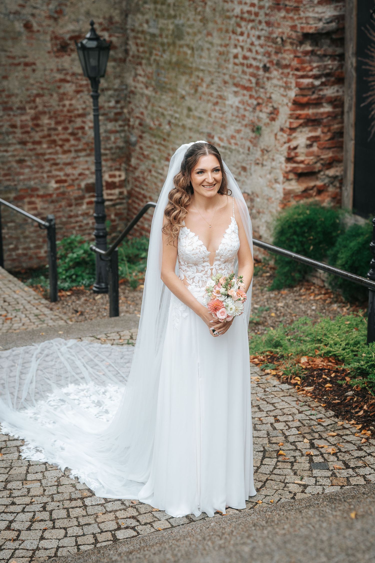Smiling bride in a lace wedding gown with a long veil holding a pink bouquet on a cobblestone path by an old brick wall.