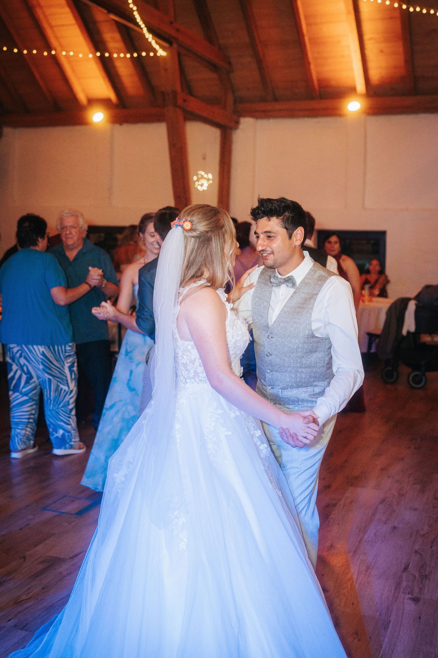 Bride in a white lace gown and veil dances with a groom in a gray vest and bow tie while wedding guests celebrate in a rustic wooden venue.