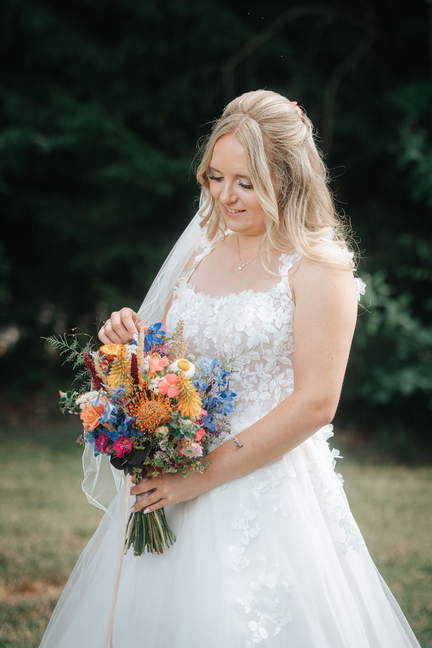 Smiling bride in a lace wedding gown and veil holds a colorful wildflower bouquet of orange, blue and pink blooms outdoors.