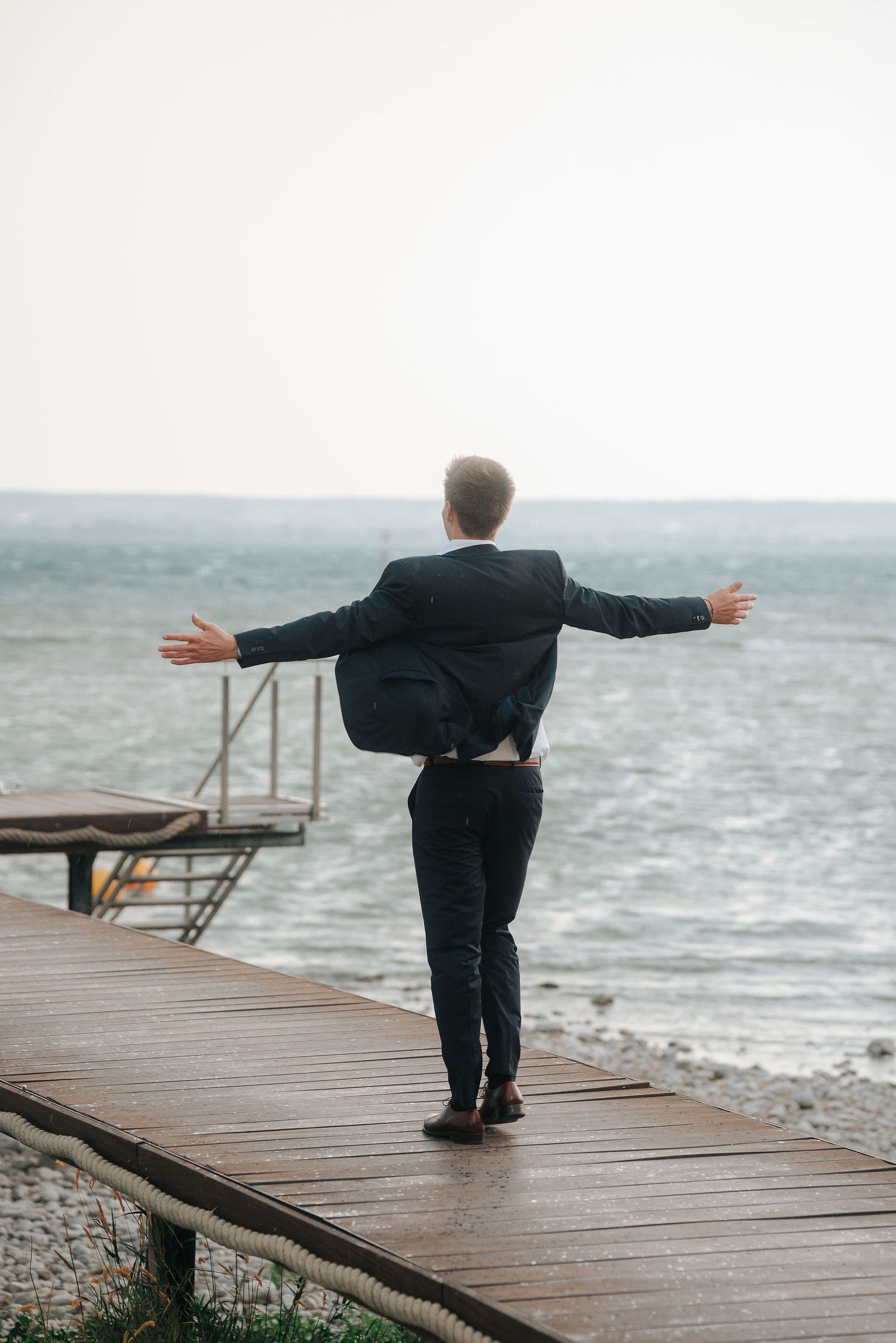 Man in a dark suit stands on a wooden pier with arms outstretched, facing the windy sea under a hazy overcast sky.