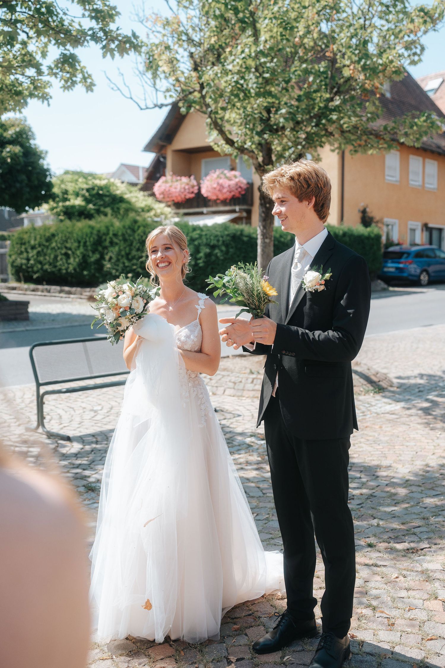 Smiling bride in a white tulle gown and groom in a black suit hold bouquets outdoors on a cobblestone street at their wedding.