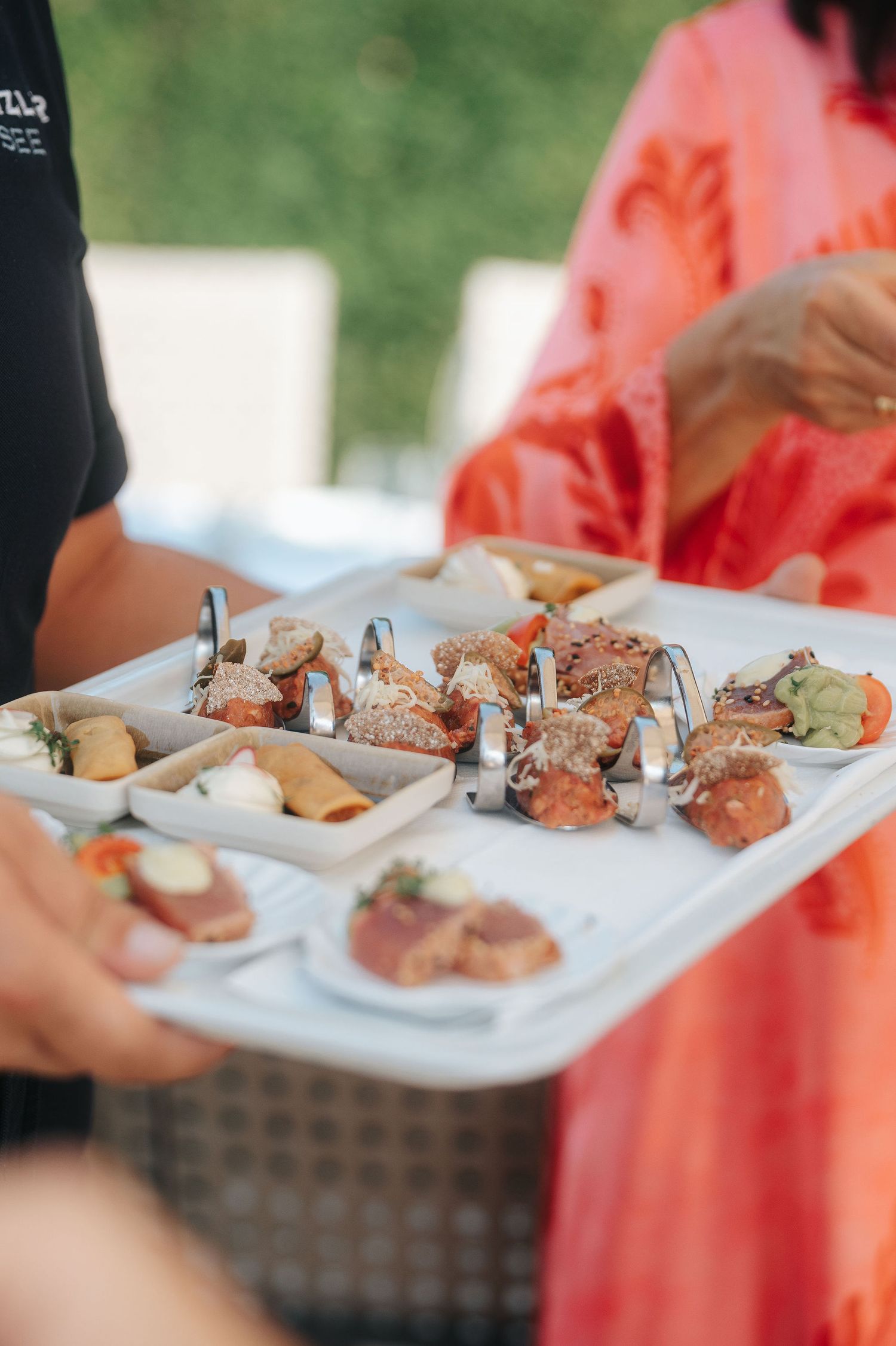 Server holding a white tray of assorted gourmet canapés and appetizers on small plates and metal holders at an outdoor catered event.