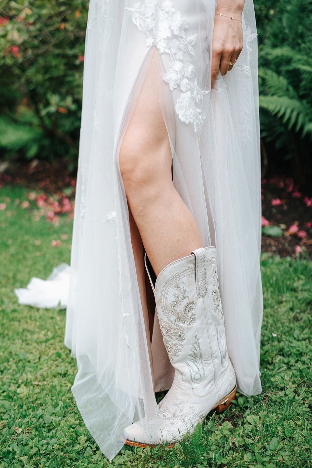 A close-up view of white embroidered cowboy boots peeking out from under the hem of a flowing wedding dress.