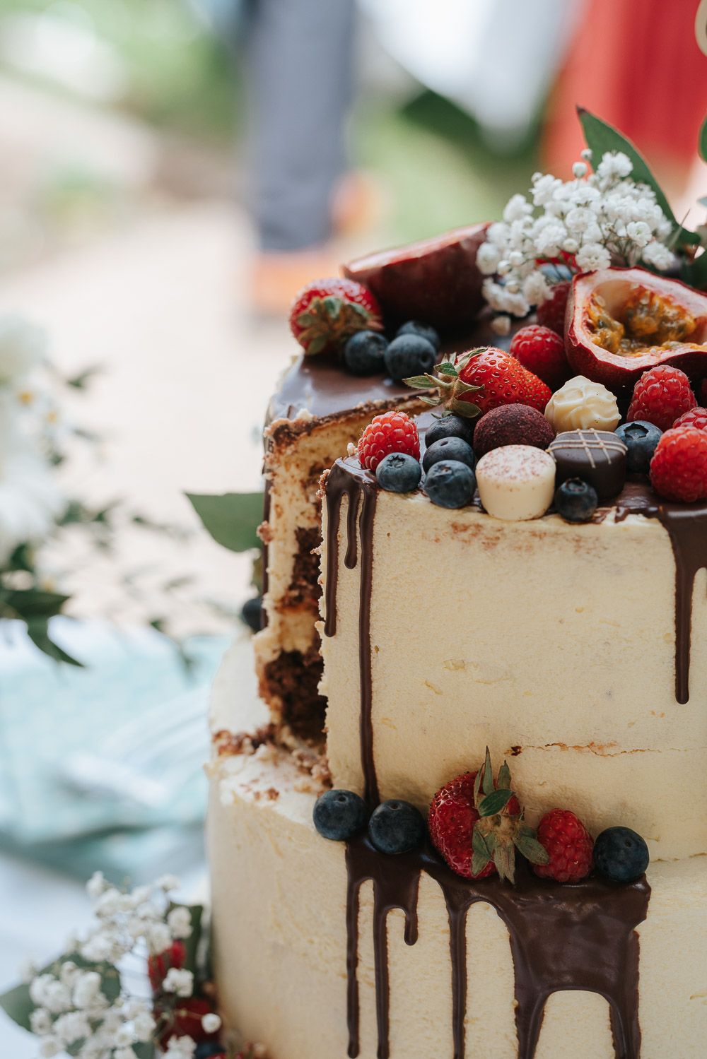 Two-tier white wedding cake decorated with chocolate drips, fresh berries, baby's breath flowers and passion fruit.