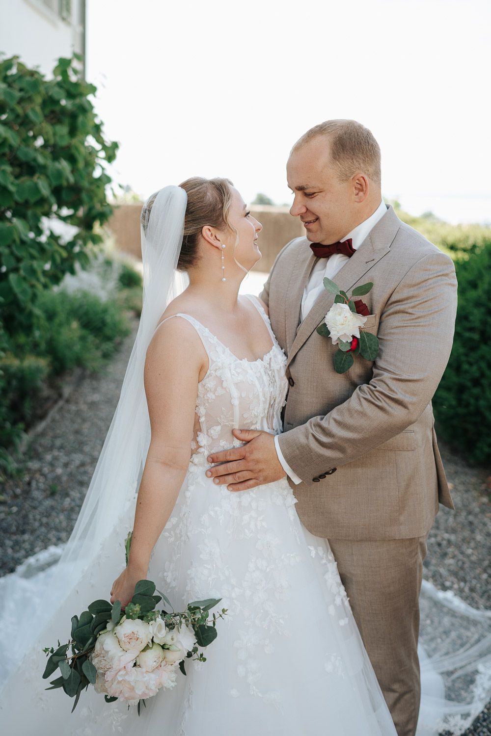 Romantic outdoor wedding portrait featuring a couple in tan suit and white lace dress with floral bouquet among greenery.