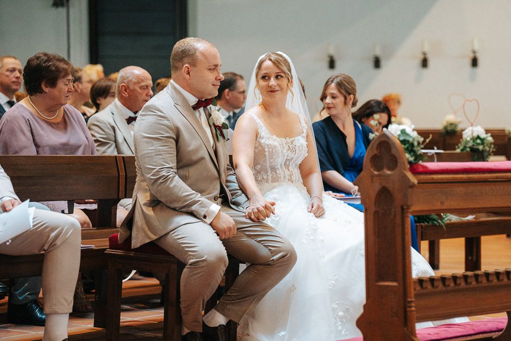 A couple sits together in wooden church pews during a wedding ceremony, with guests visible in the background.