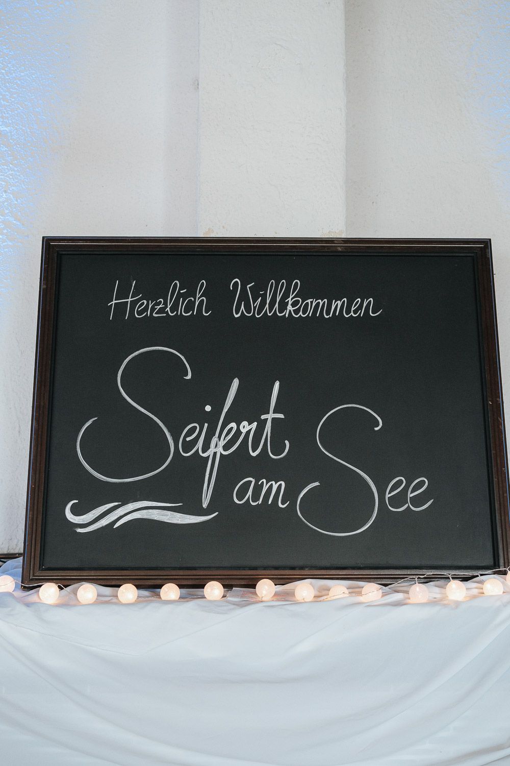 A black chalkboard sign displaying 'Herzlich Willkommen Seifert am See' in white cursive writing with string lights below.