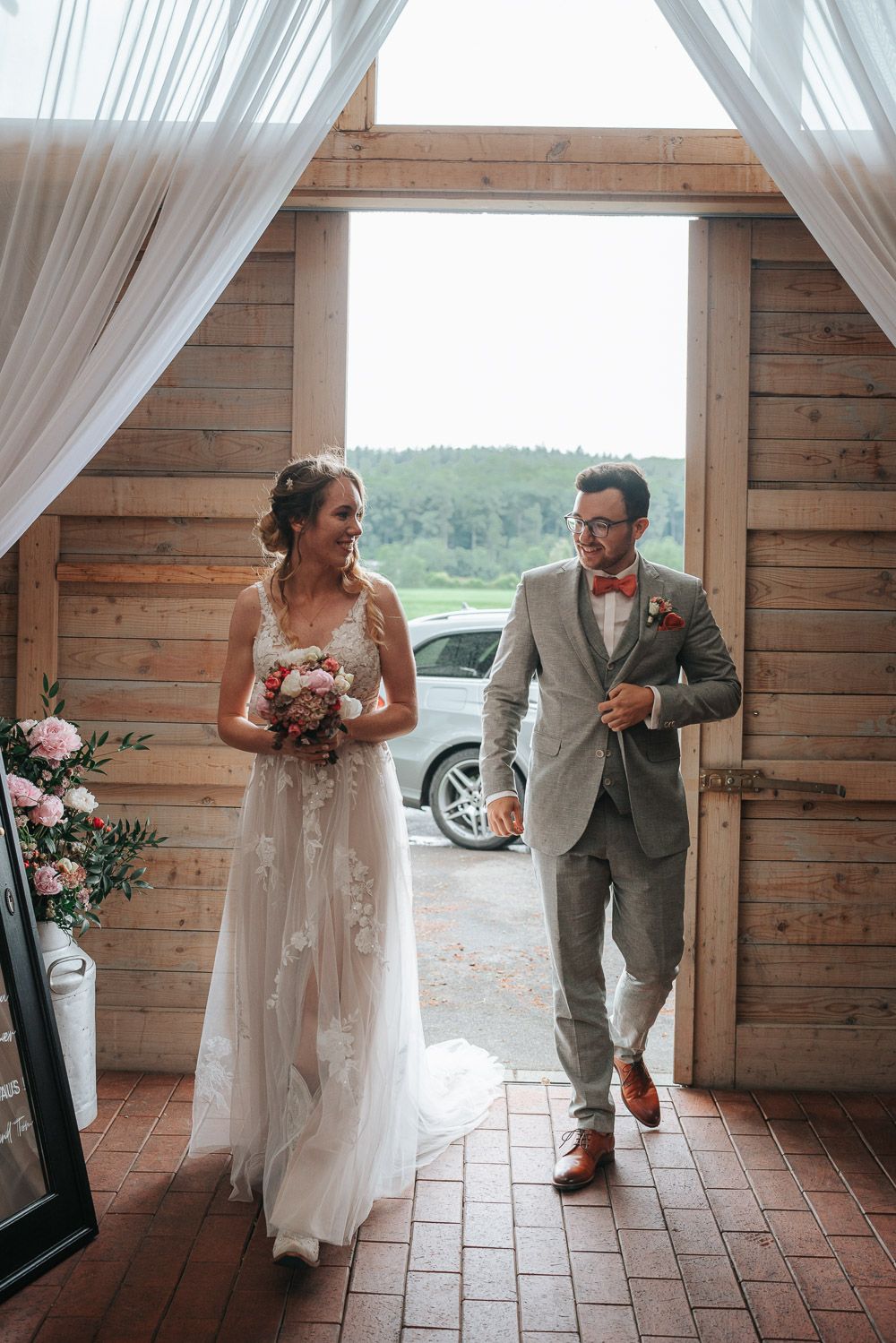 Wedding party members playfully run and dance along a covered brick porch with white draped curtains.