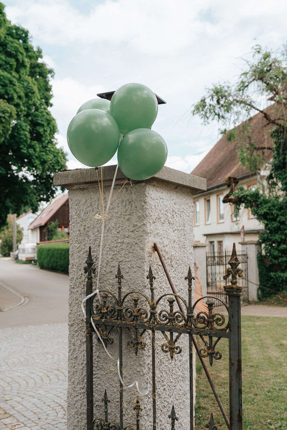 Three mint green balloons tied to an ornate metal gate post outside a residential house on a quiet suburban street.