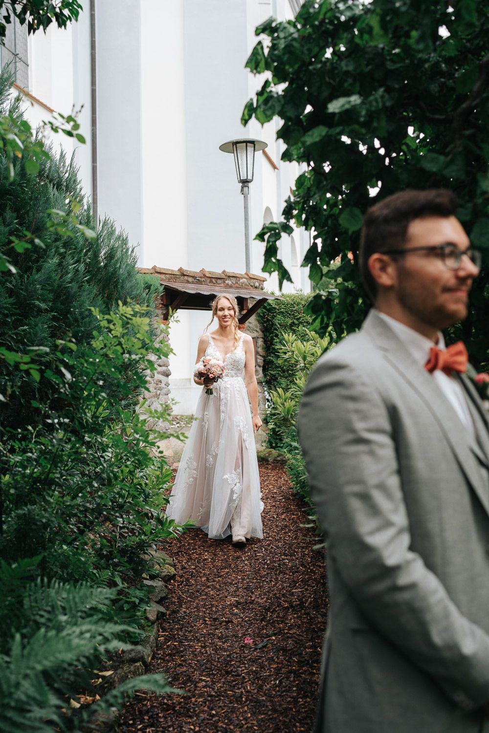 A first look moment at a wedding with the bride in a flowing lace gown walking down a garden path.